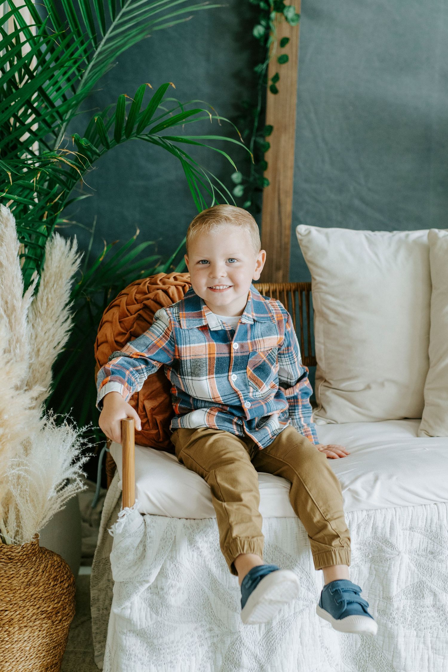 Child sits on white sofa with plaid shirt and khaki pants surrounded by tropical plants and boho decor.