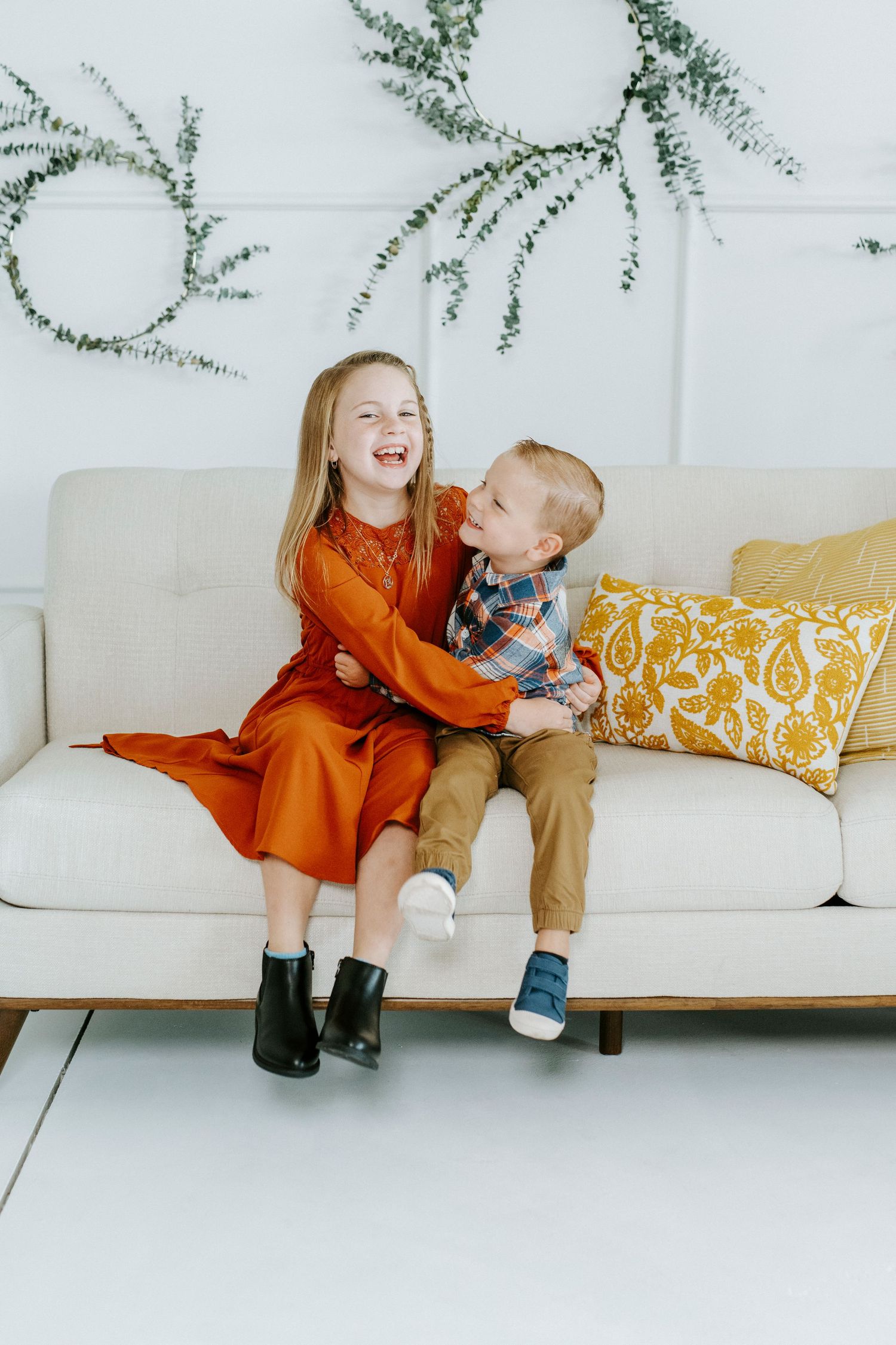 Two children sitting and laughing together on a white couch with decorative greenery hanging on the wall behind them.