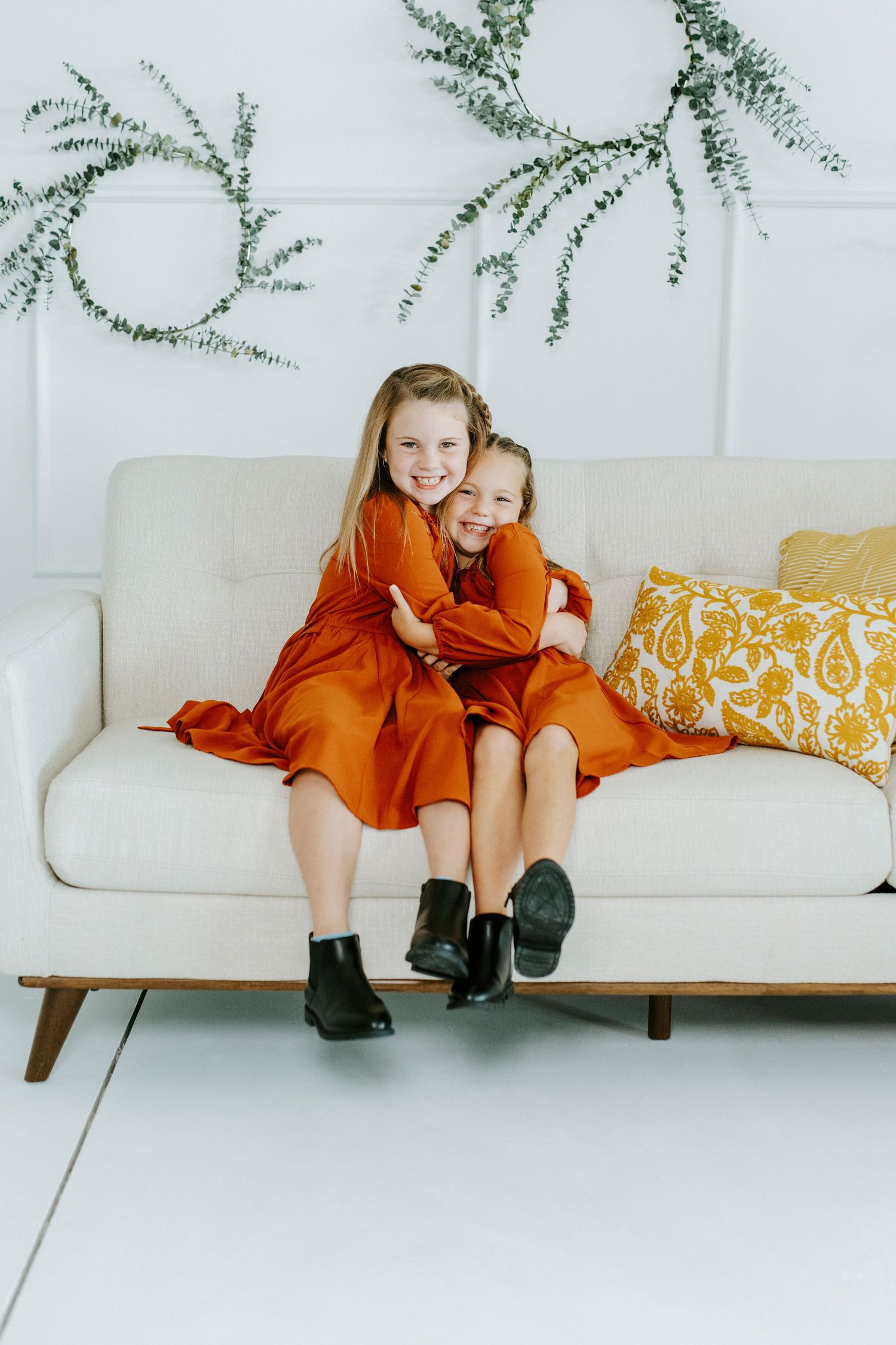 Two children in matching burnt orange dresses sit together on a modern white couch with botanical wall decor.