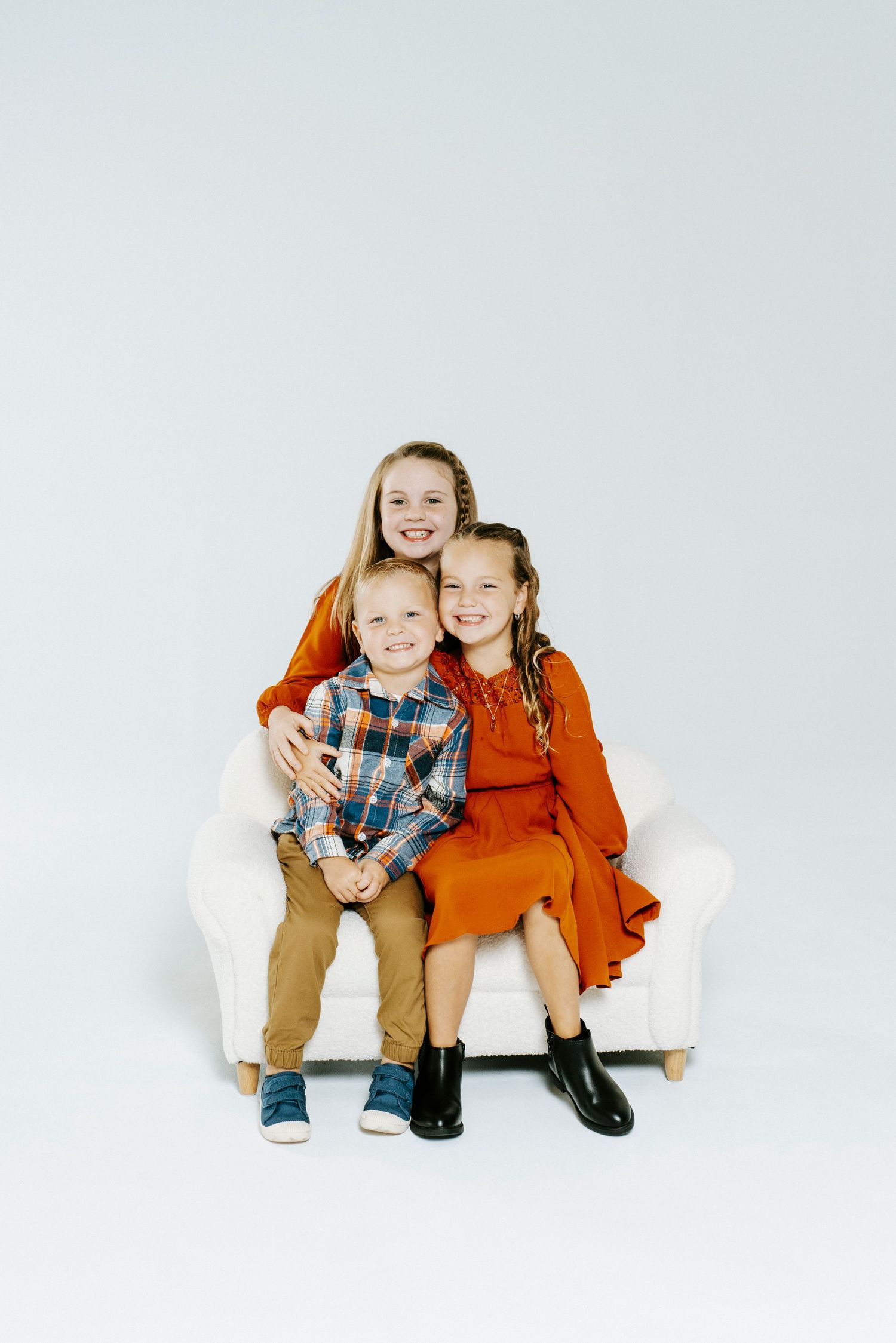 Three children sitting together and smiling on a white couch against a plain background.
