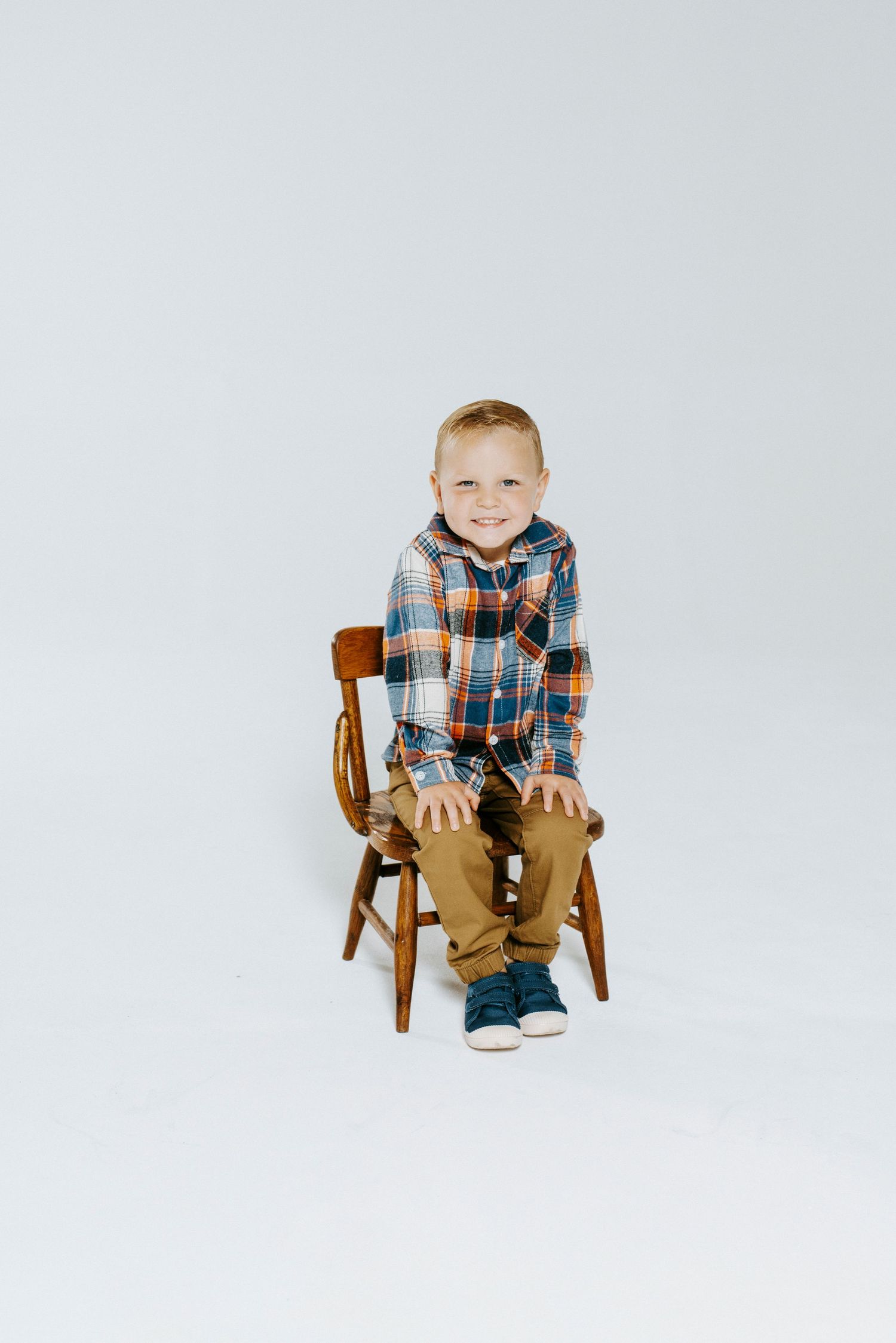 A minimalist photo series shows a young child in plaid sitting on a wooden chair against a white backdrop.