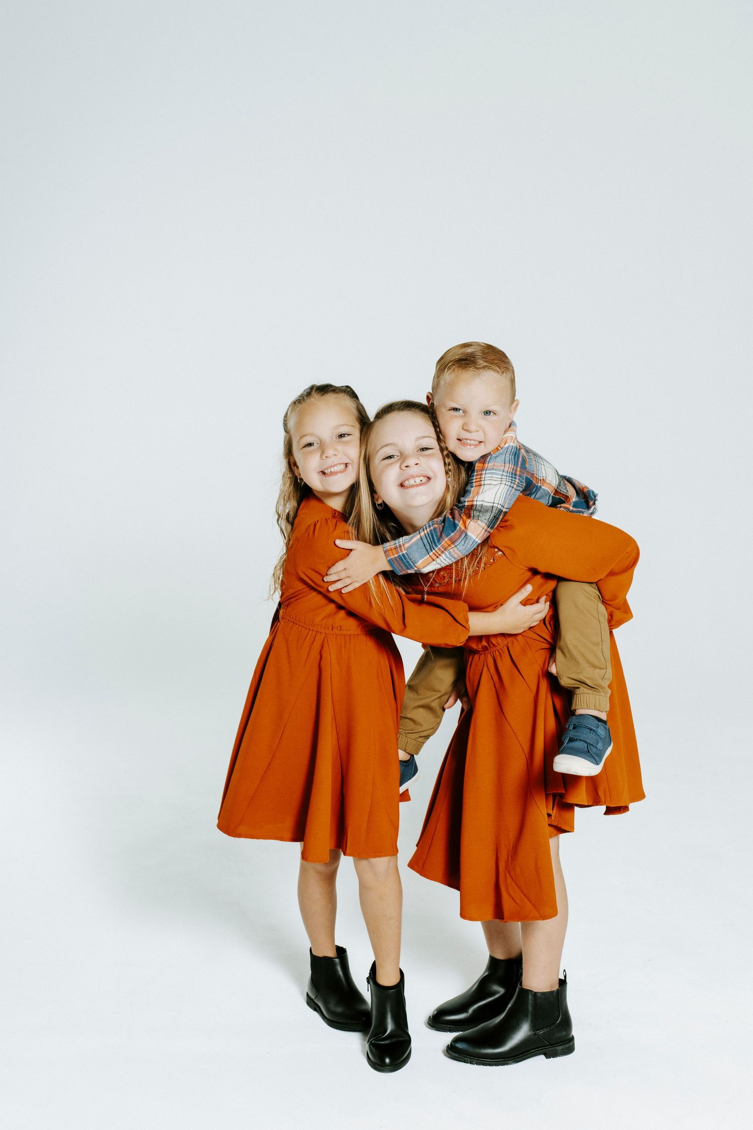 Three young children wearing orange dresses and dark shoes pose playfully together against a light background.