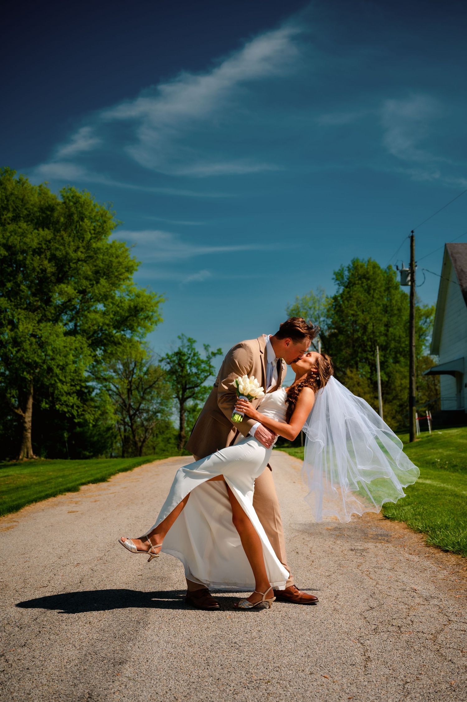 Bride and groom share a romantic dip kiss on a sunny country road, with blue skies and green trees behind them.