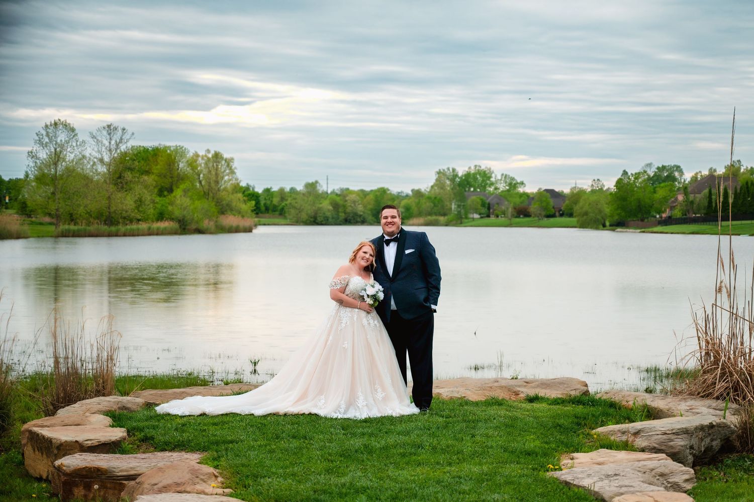 A couple in wedding attire stands by a serene lake with lush green grass and trees in the background.