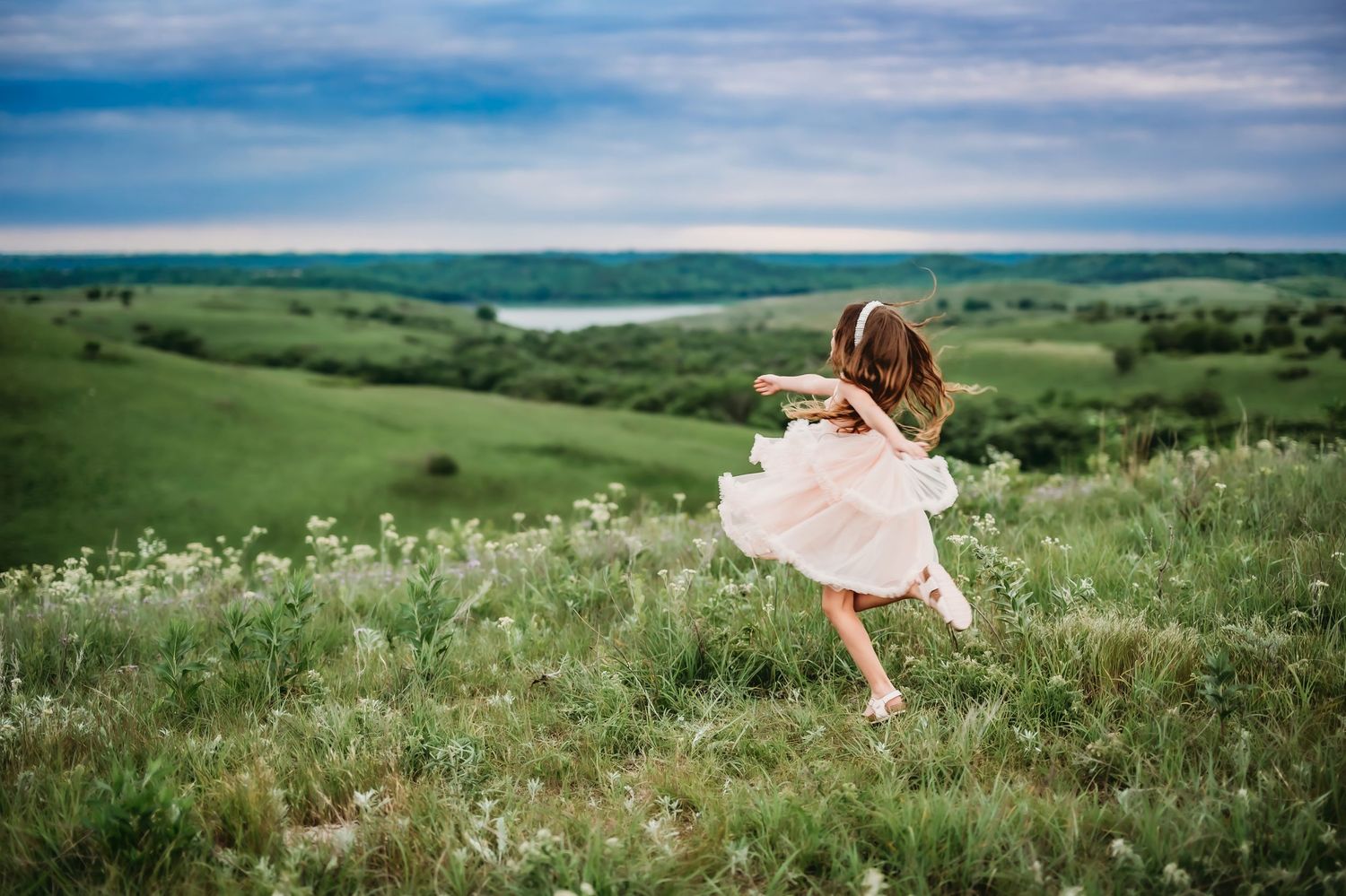 A dancer in white dress twirls gracefully on a hilltop meadow overlooking a scenic green valley landscape.