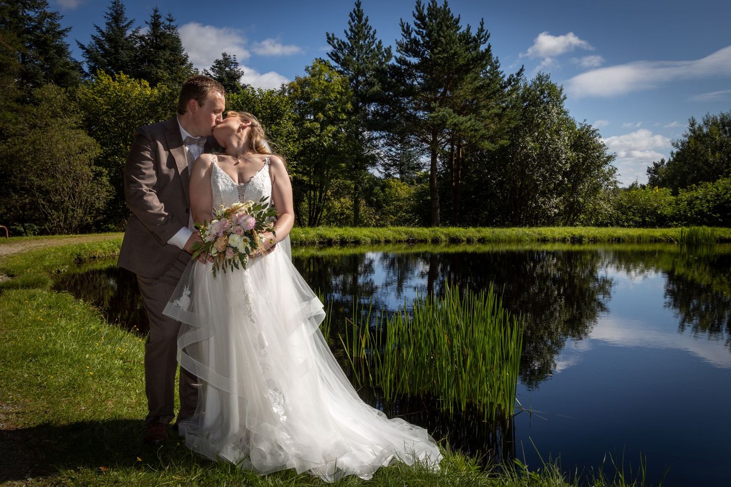 Bride and groom kiss romantically beside serene reflective pond surrounded by lush greenery.