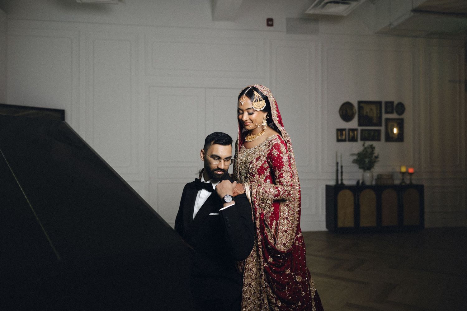 A bride in a red sequined lehenga and groom in black suit pose together in modern white architecture.