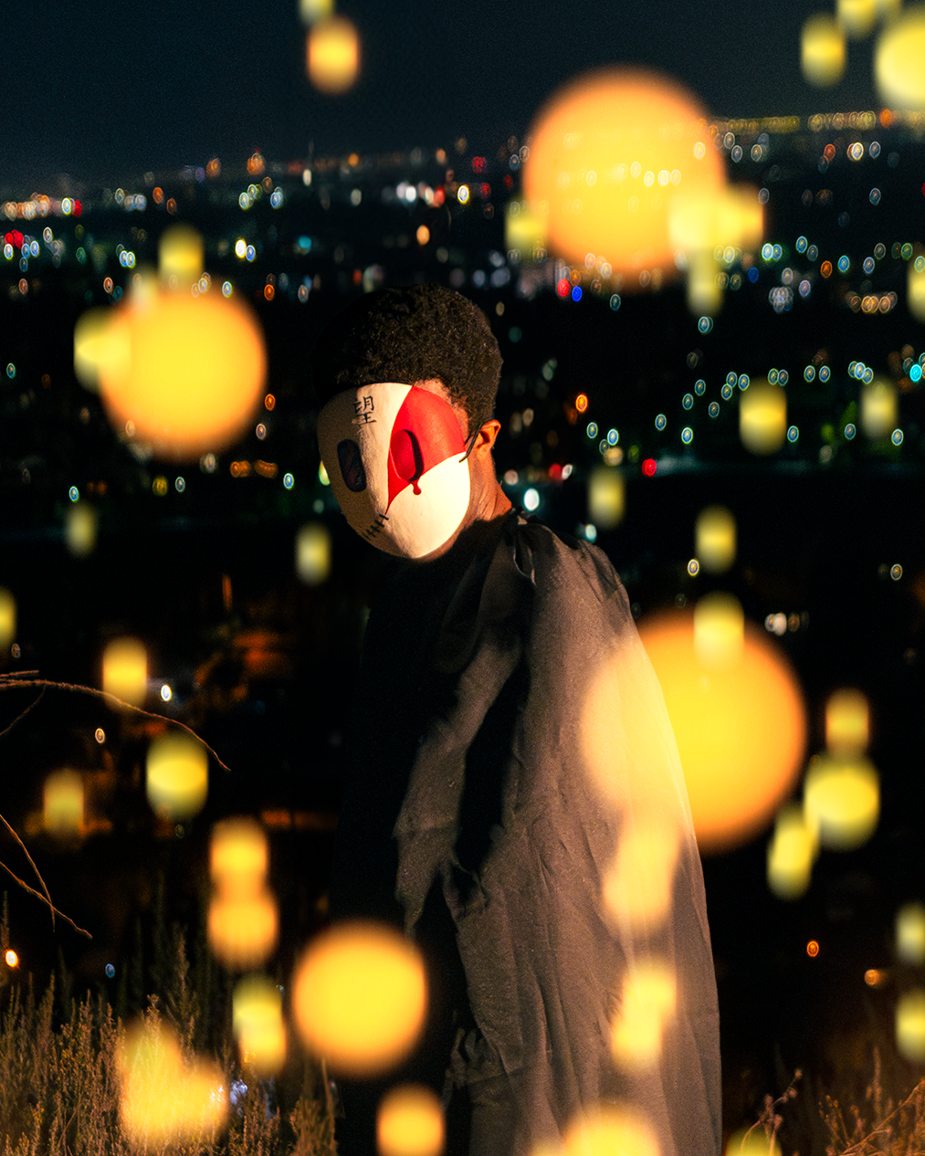 A masked character stands against the backdrop of a bokeh city nightscape. Balls of bright orange-yellow lights float up around him.