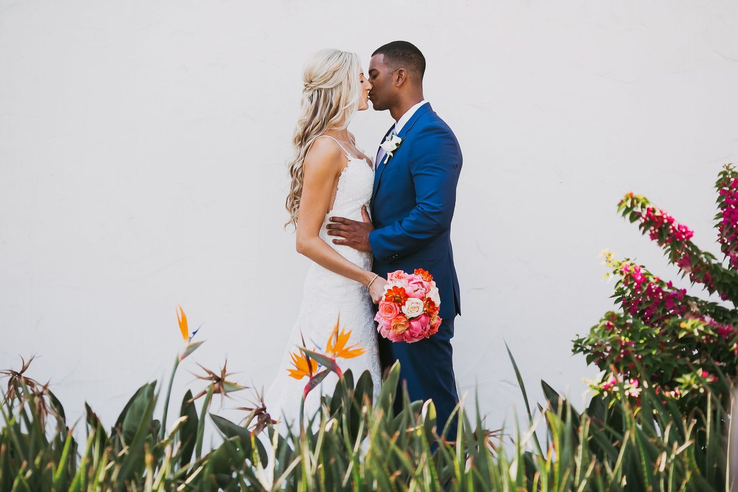 Couple walking through colorful garden with tropical flowers and white wall background.