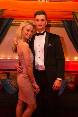 A couple poses together at a formal event with red lighting and elegant decor in the background.