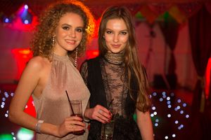 Two women in formal attire smile while holding drinks at an evening event with red lighting and blue lights in background.