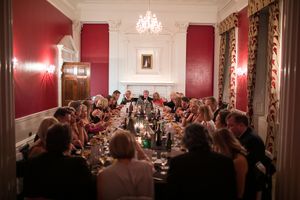 A formal dining room with red walls and crystal chandeliers hosts a crowded dinner event.