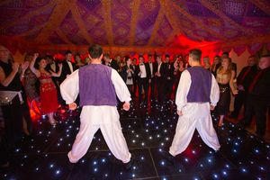 Dancers in white and purple costumes perform a traditional folk dance routine on a stage with blue lighting.
