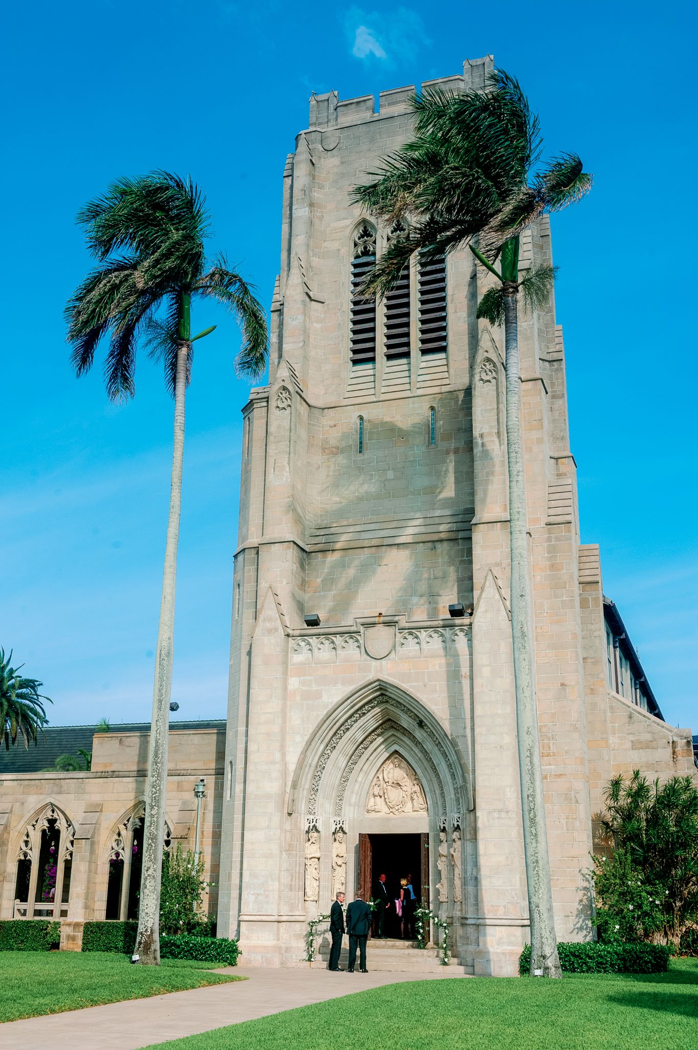 A Gothic style church with tall stone walls and palm trees swaying against a bright blue sky in Palm Beach Florida.