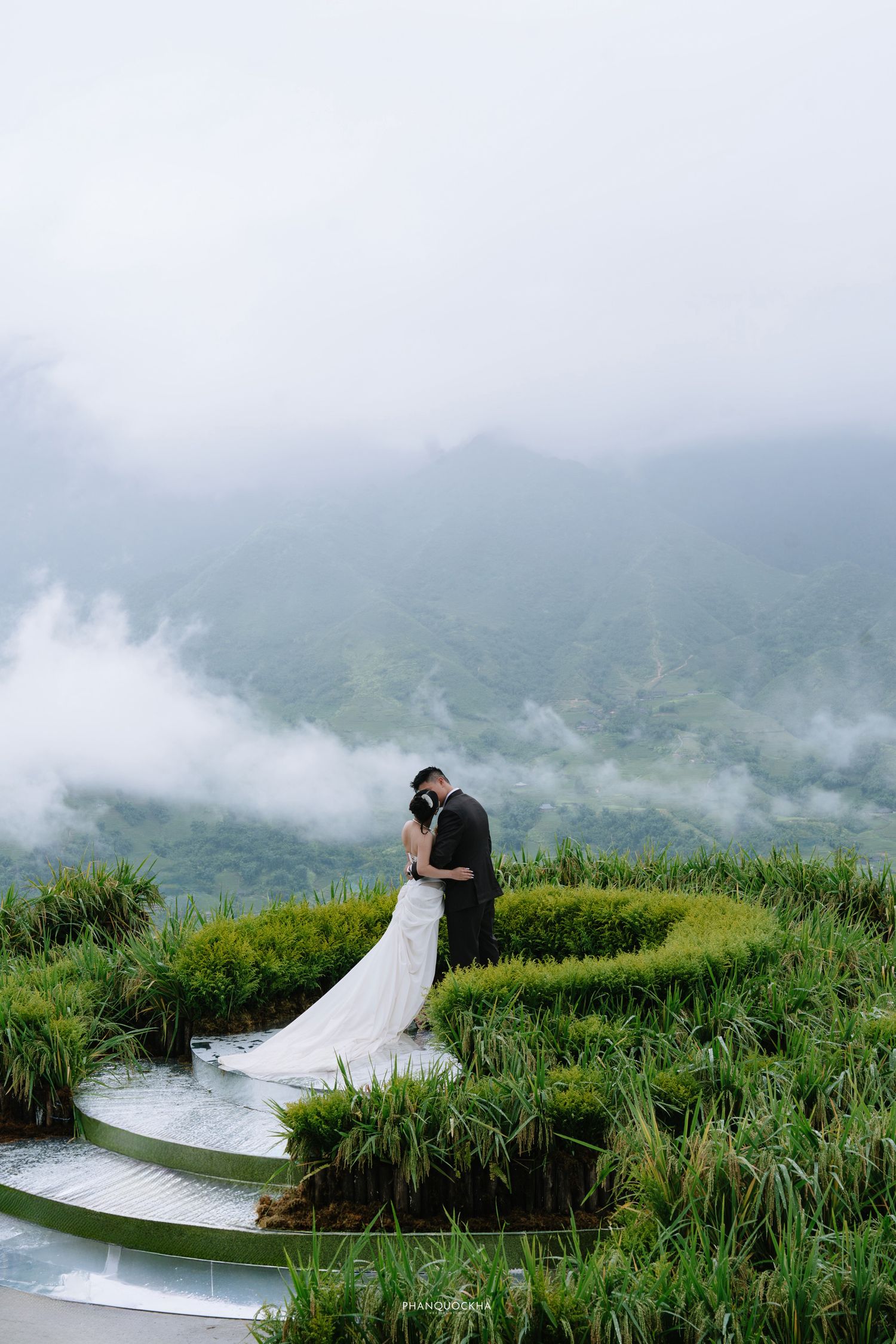 Bride and groom share intimate embrace on spiral rice terrace with foggy mountain scenery behind.