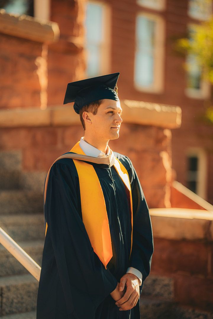Graduate in academic regalia with gold stole poses at golden hour near brick building.