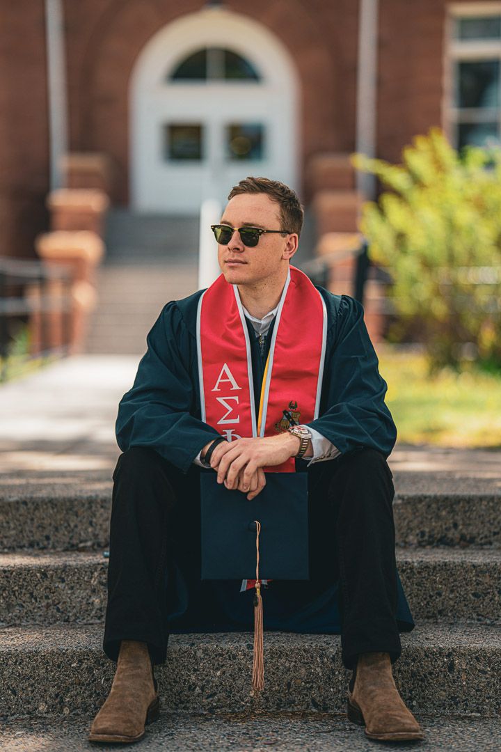 Graduate in navy cap and gown with sunglasses and red stole sits contemplatively on concrete steps.