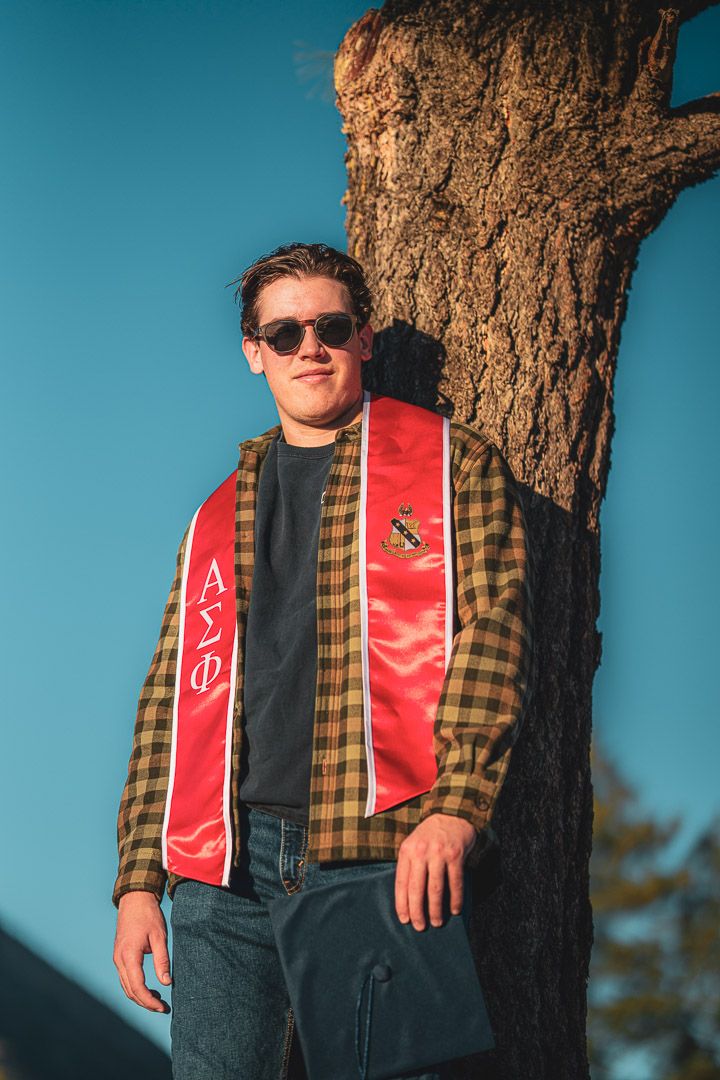 Student wearing sunglasses and red fraternity stole poses against large tree trunk.