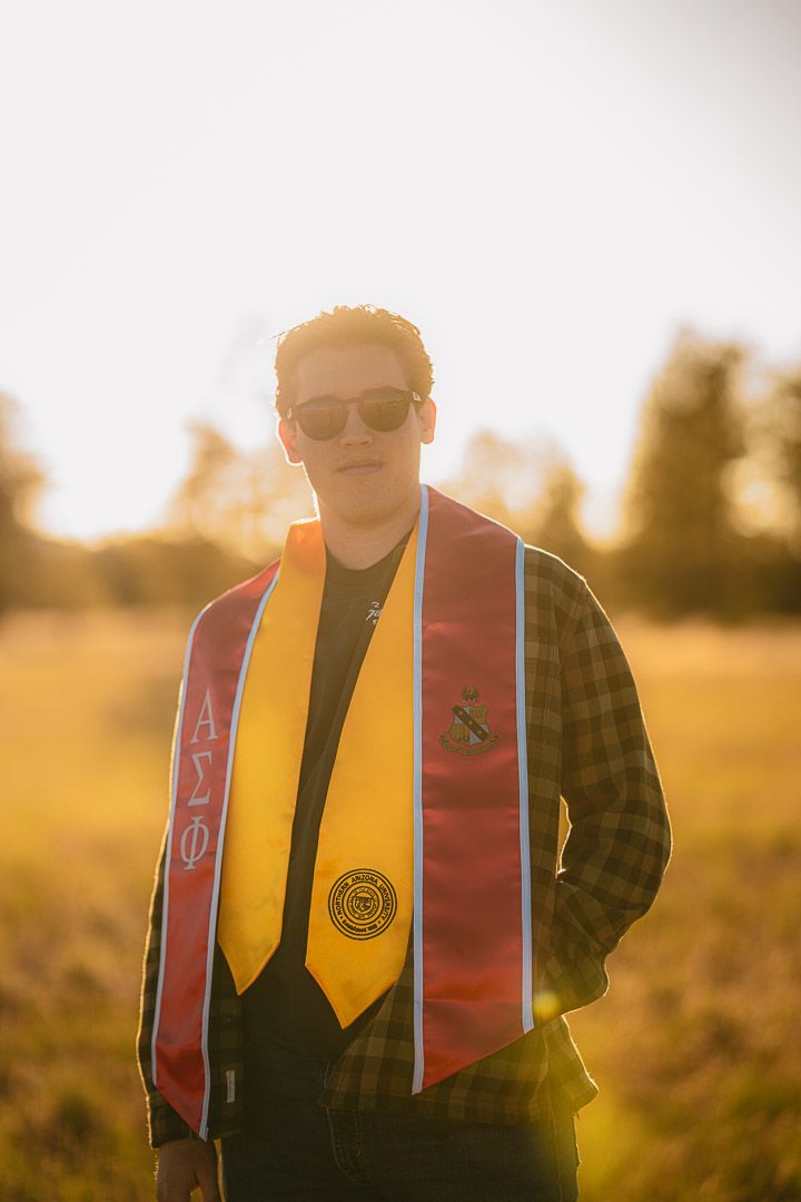 Backlit portrait at golden hour showing someone wearing graduation stoles and sunglasses.