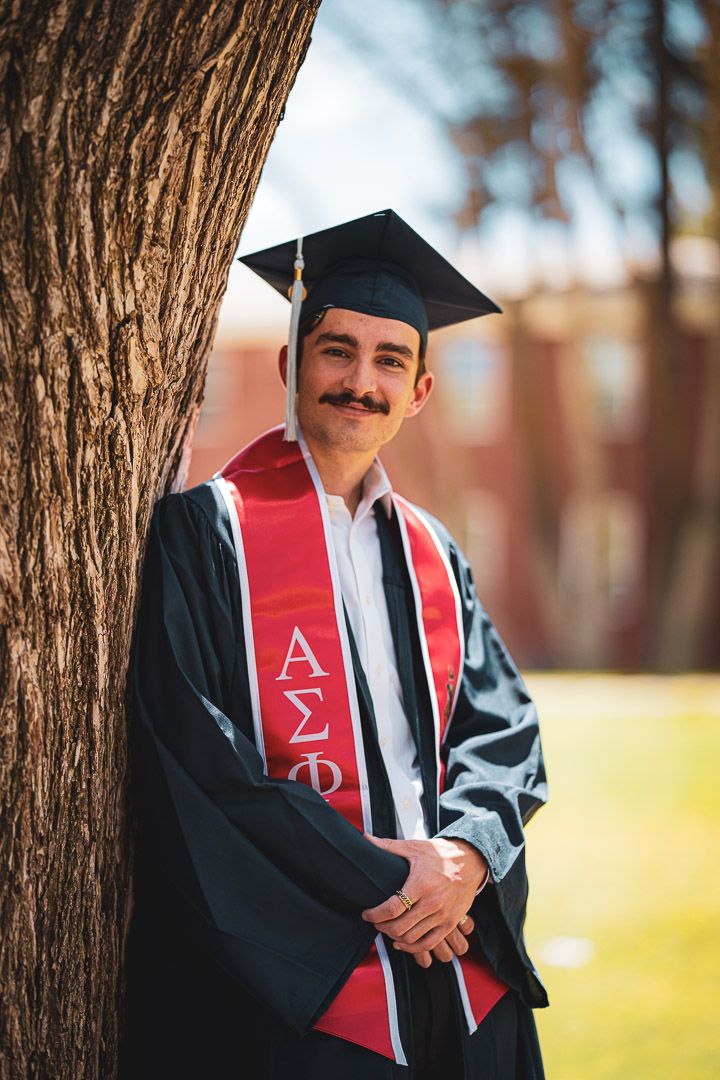 Graduate in black cap and gown with red fraternity sash poses beside large tree trunk.