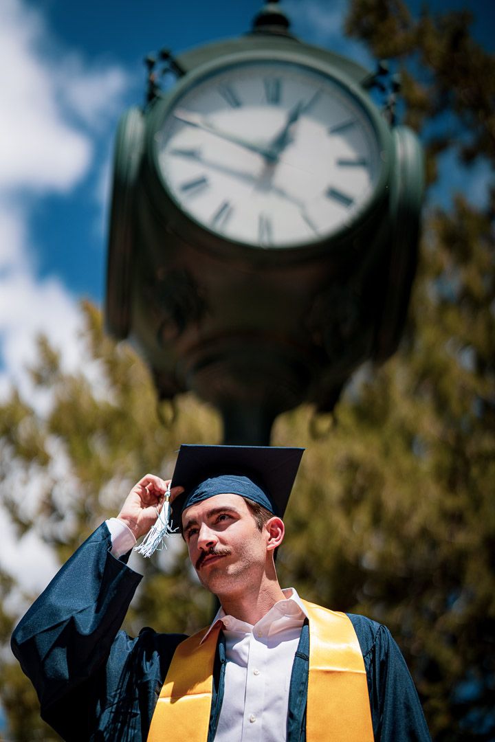 Graduate in cap and gown poses beneath vintage clock tower during graduation ceremony.