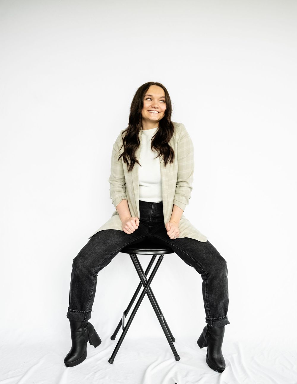 Person in blazer and dark pants poses on black folding chair against white background.