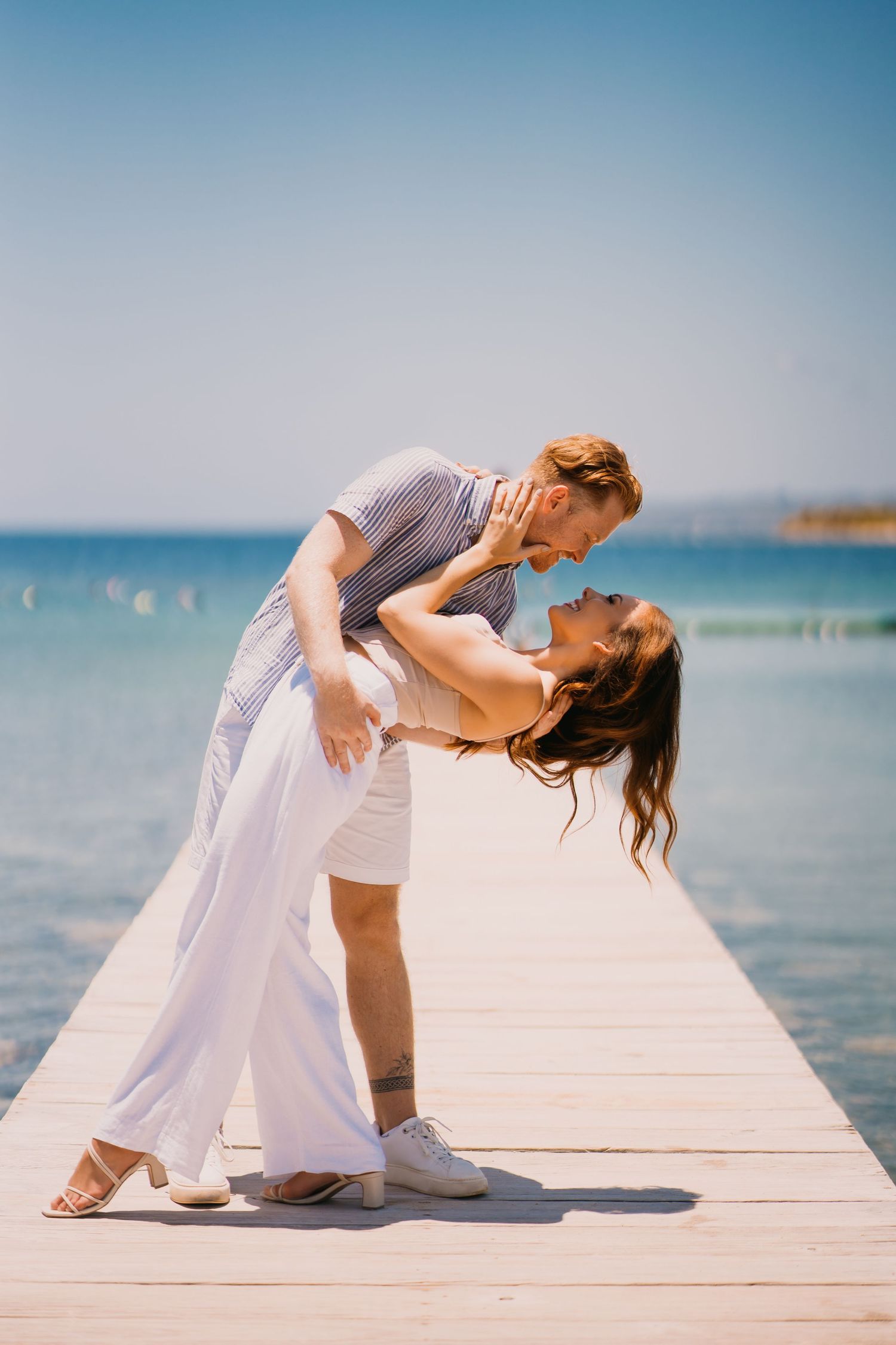 A romantic couple in white clothing dances gracefully on a wooden pier overlooking the blue ocean.