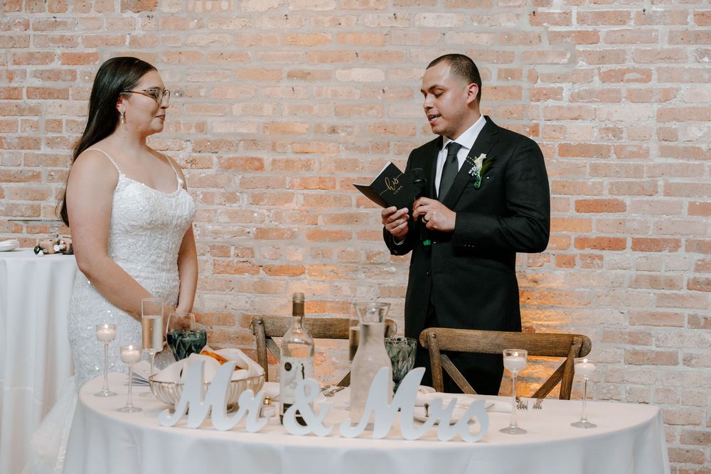 Two people share a moment at an elegant wedding reception table with decorative place settings against a brick wall backdrop.