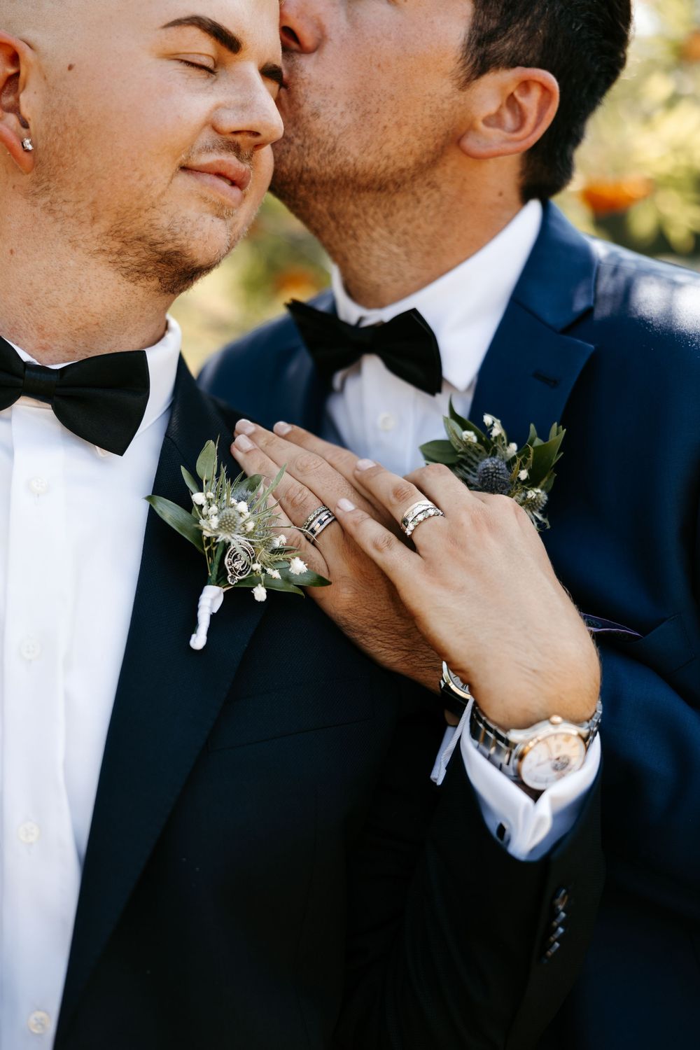 Close up photo of wedding couple in black tuxedos showing rings and boutonniere with greenery.