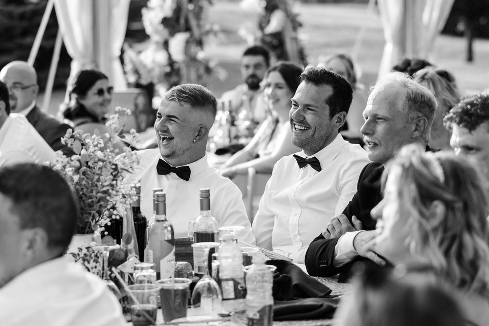 Wedding guests laugh and smile together at an outdoor reception table in this black and white photograph.