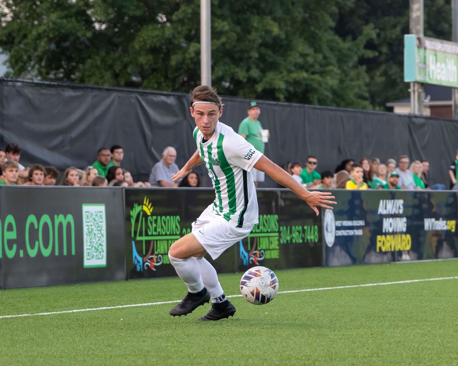 Soccer player in green and white striped jersey controls ball during match with spectators watching from sideline.
