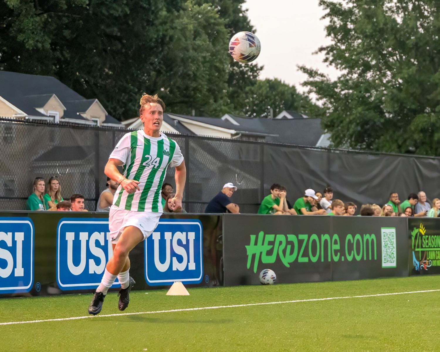 Soccer players in green and white striped uniforms compete on a grass field with advertising boards visible in background.