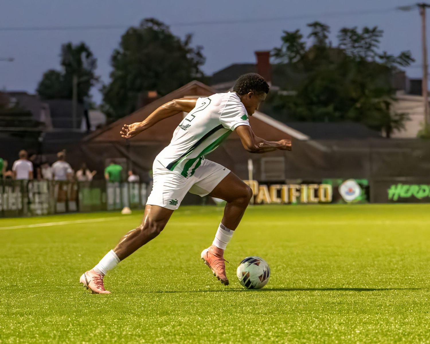 Soccer player in white uniform dribbles the ball on illuminated grass field during evening match.