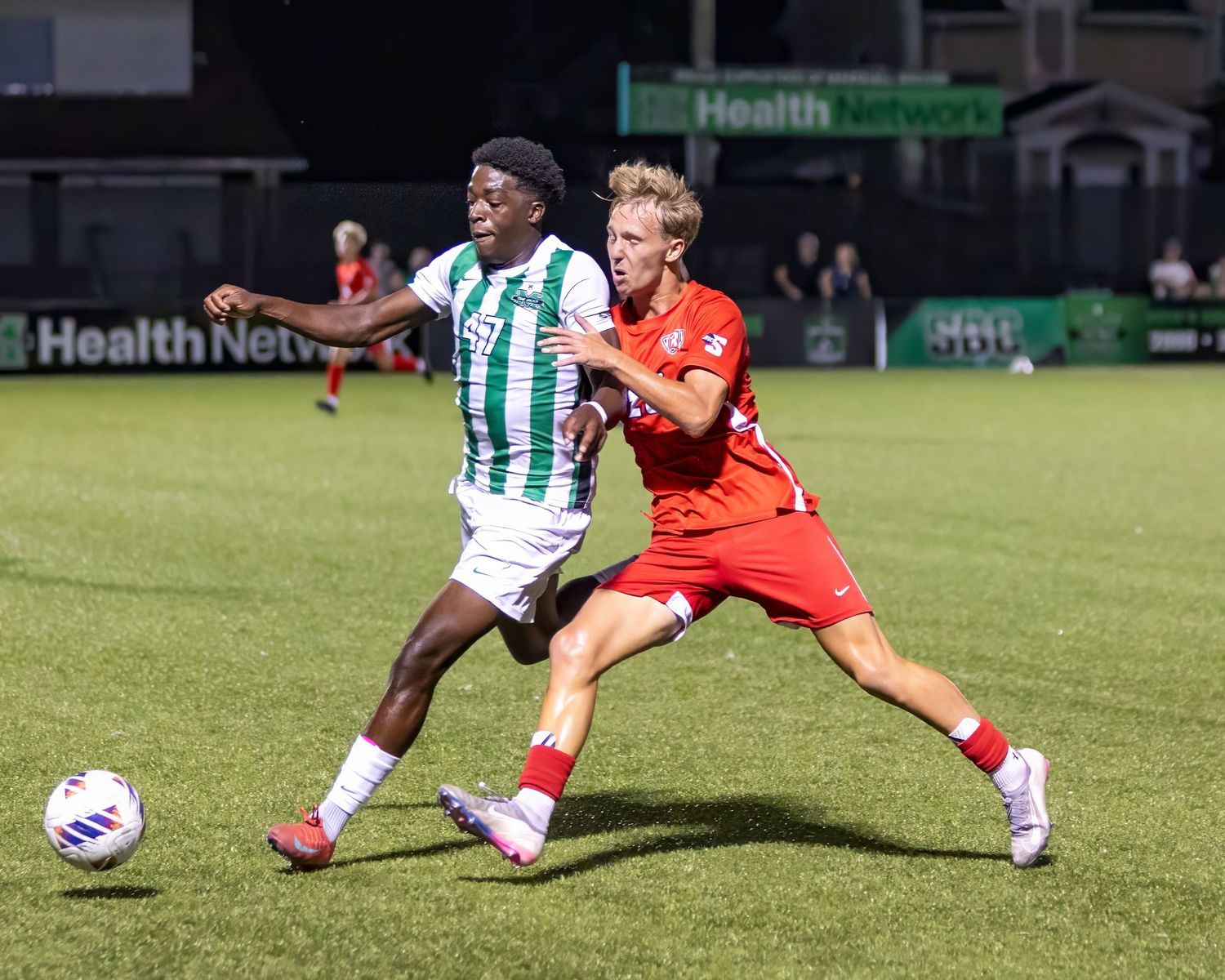 Soccer players in green-white striped and orange jerseys compete for the ball during a nighttime match.