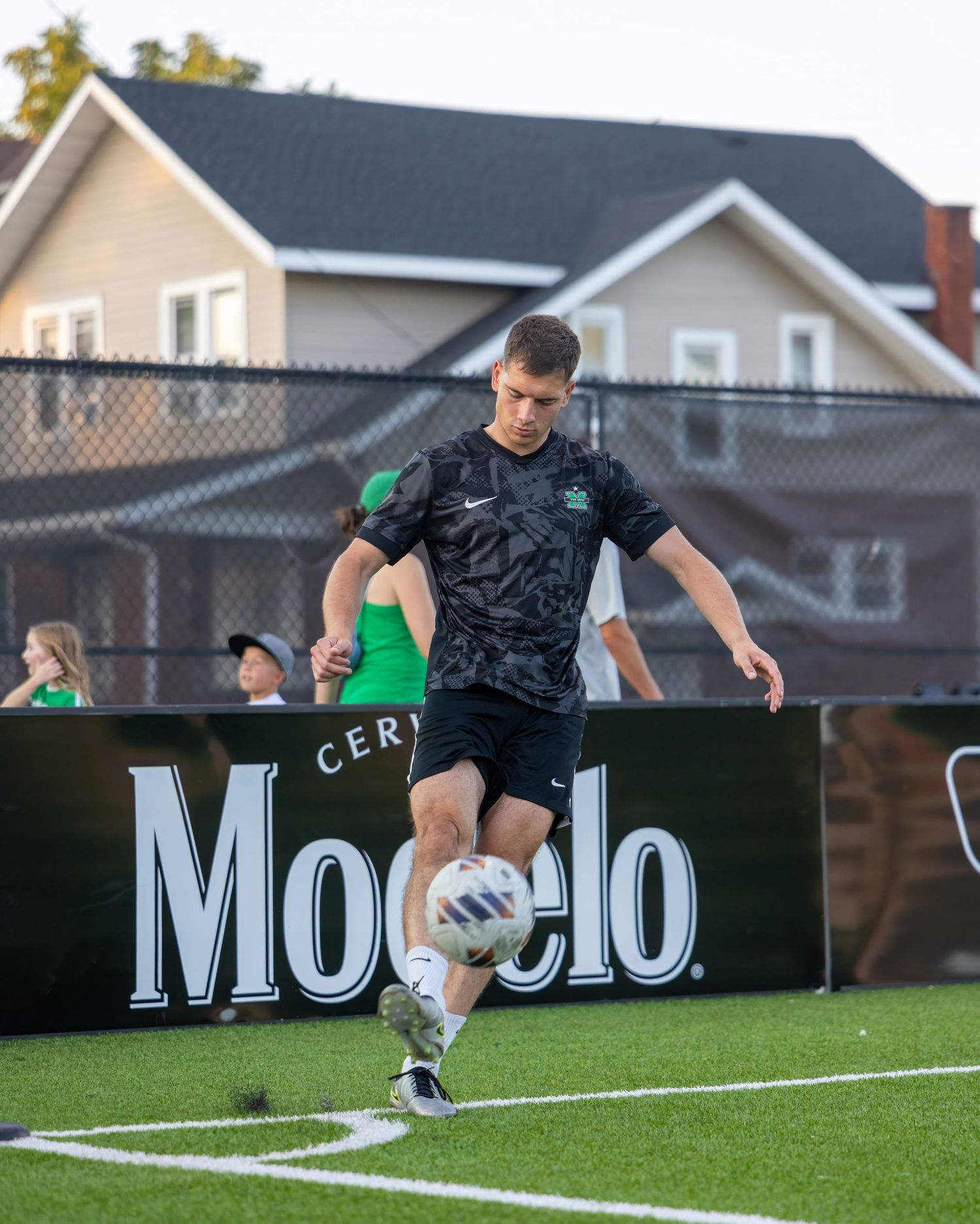 Soccer player dribbling ball on turf field next to Modelo advertisement board during match.