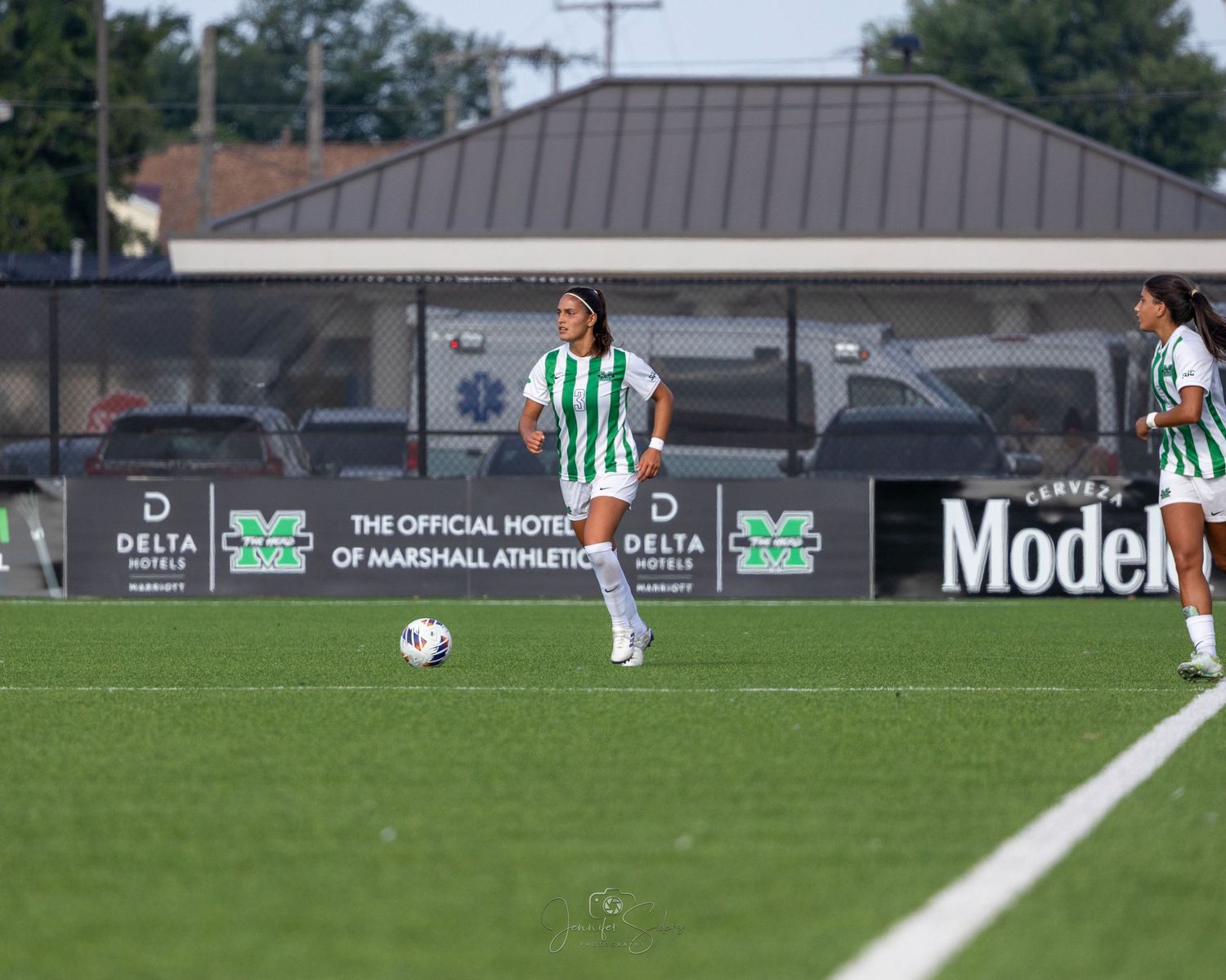 Soccer players compete on a green field with trees and buildings visible in the background during a match.