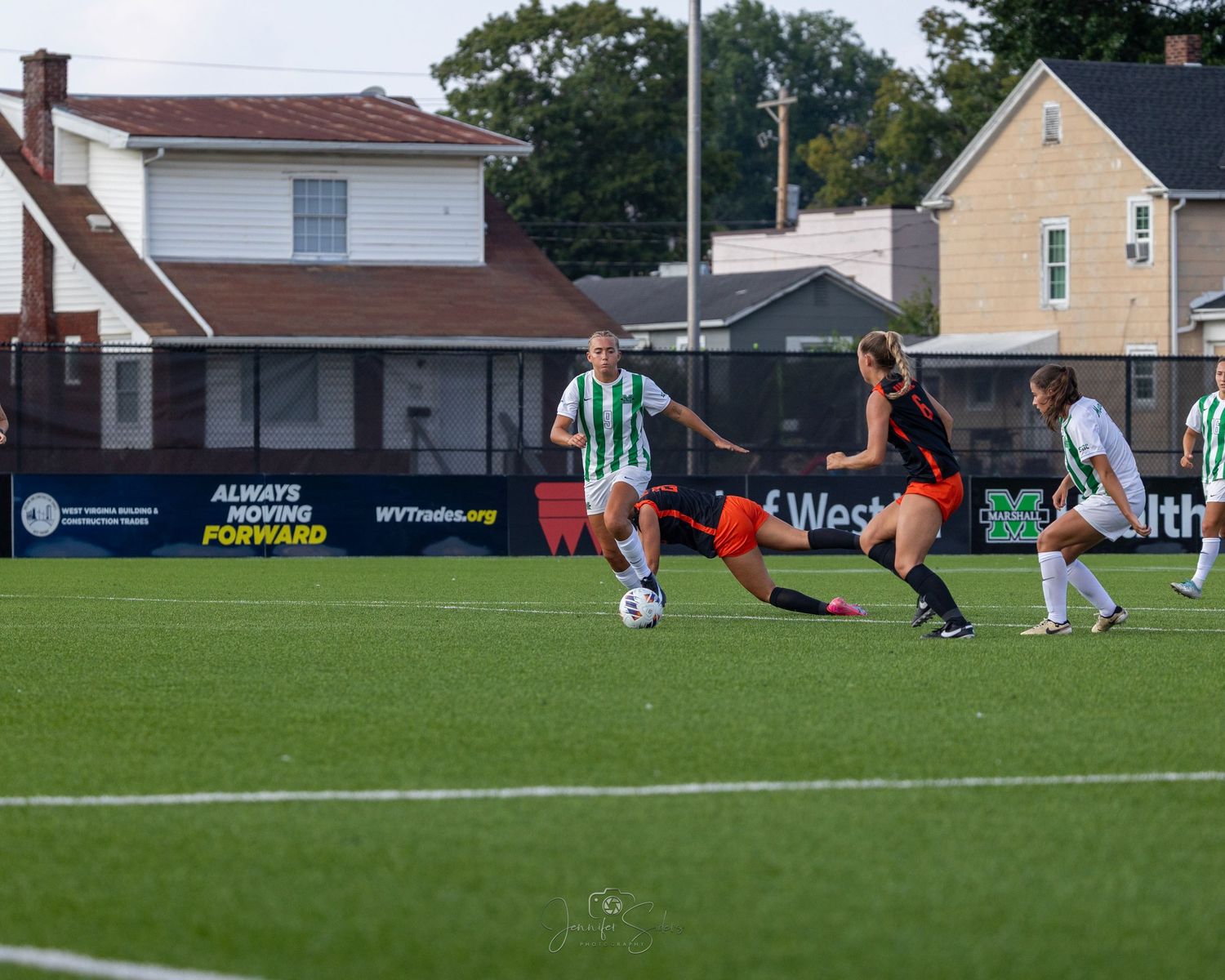 Soccer players compete for possession during a match on an artificial turf field next to residential buildings.
