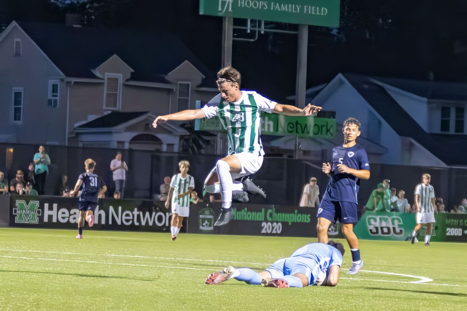Soccer player in green and white striped jersey leaps over fallen opponent during evening match on turf field.