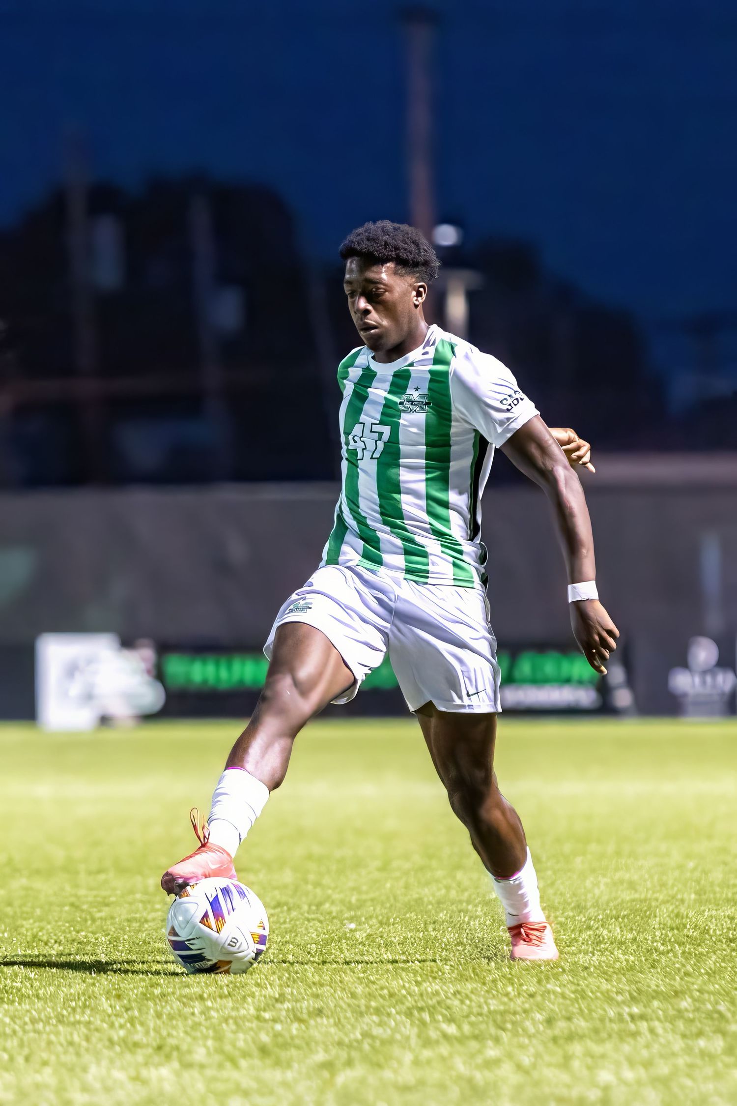 A soccer player runs on a grass field with green and white striped jersey while dribbling the ball in sunlight.