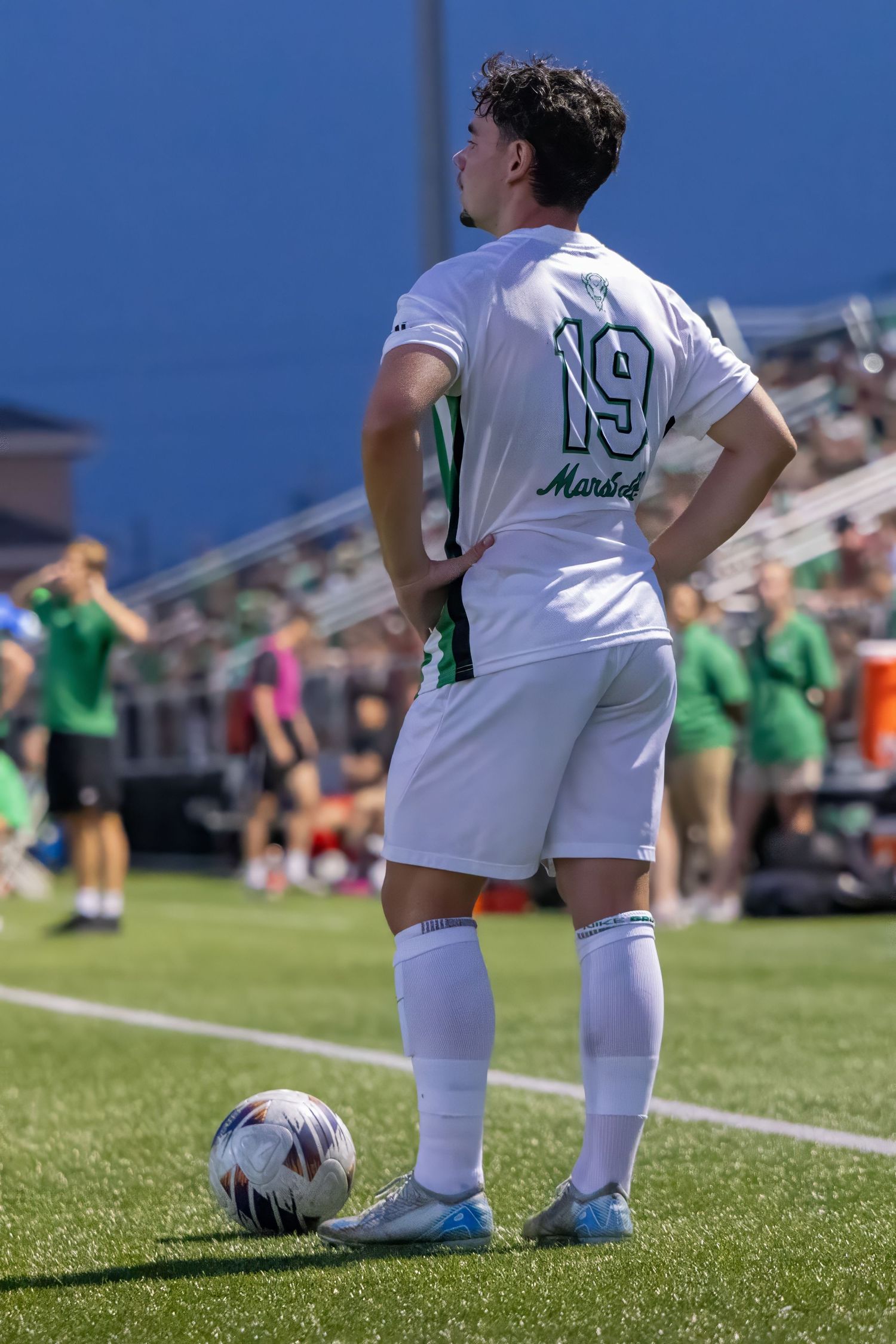 Soccer player wearing number 19 white jersey stands with ball at feet on sunny field during match.