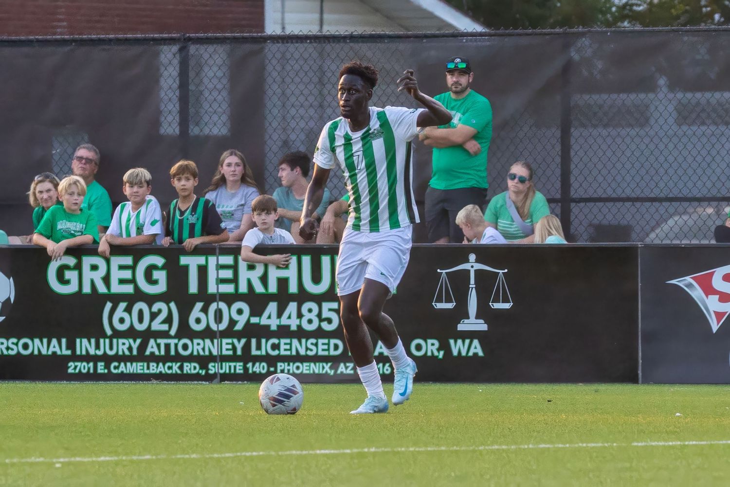 Two soccer players compete for the ball during a match with an attorney advertisement banner visible in the background.