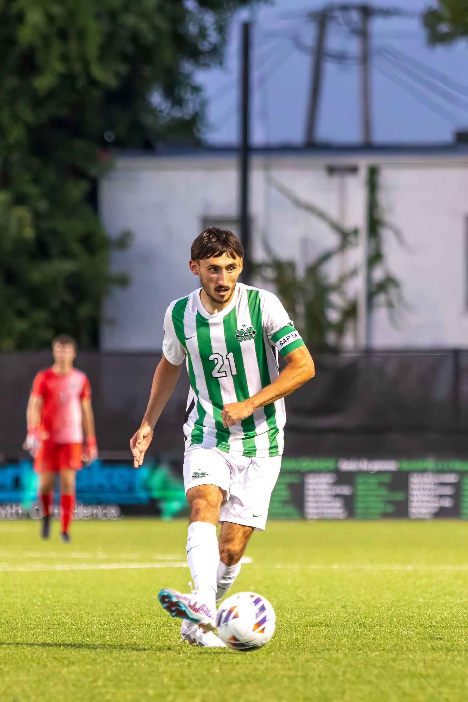 Soccer player in green and white striped jersey dribbles ball on grass field during evening match.