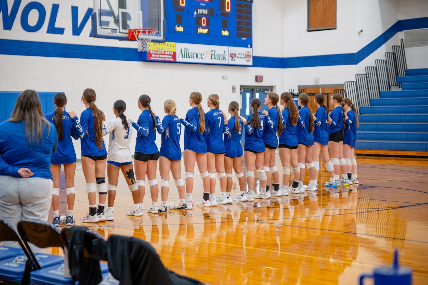 High school volleyball players in blue uniforms stand in a line during practice in a gymnasium with polished wooden floors.