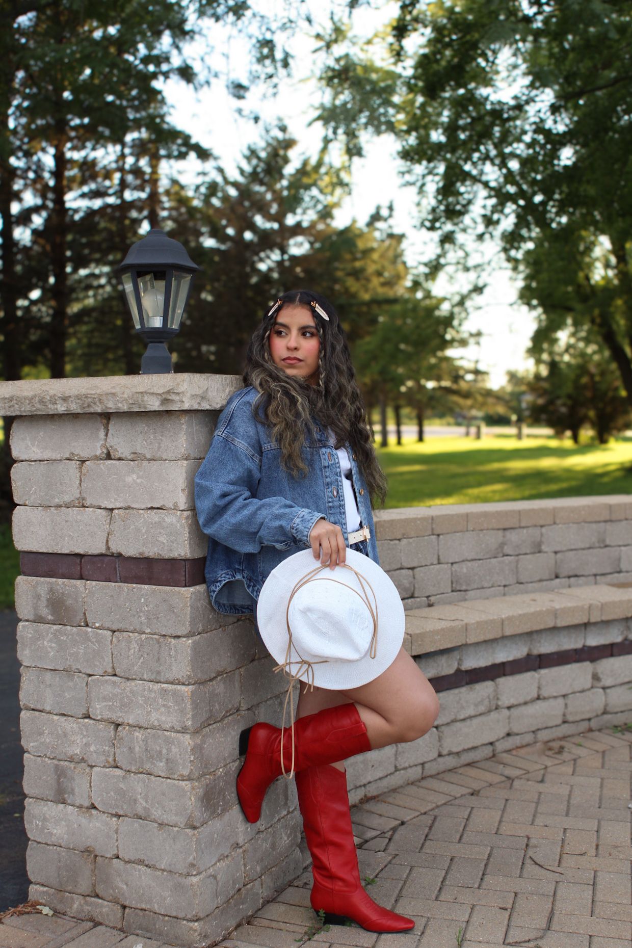 Person in denim jacket and red boots poses against brick wall with white cowboy hat.