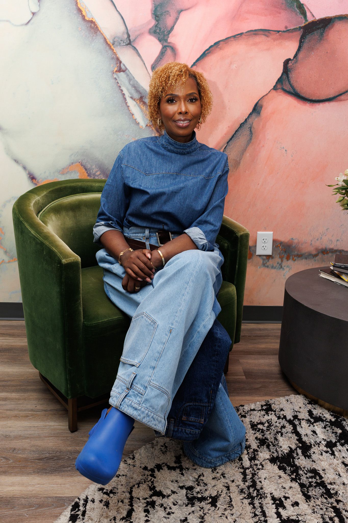 Person sitting in green velvet accent chair wearing denim jeans and blue top against pink marbled wall.