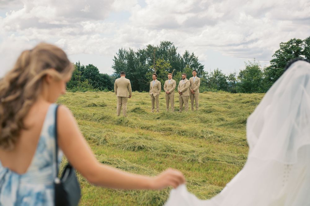 Wedding party in tan suits stands in a grassy field surrounded by pine trees under cloudy skies.