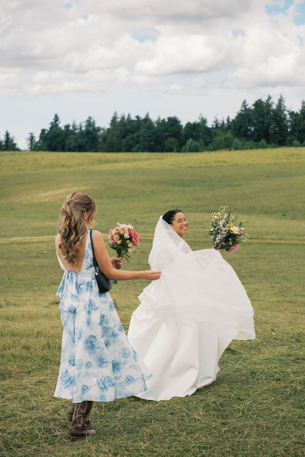 Wedding party members hold flowing white dress skirts in a sunny meadow.