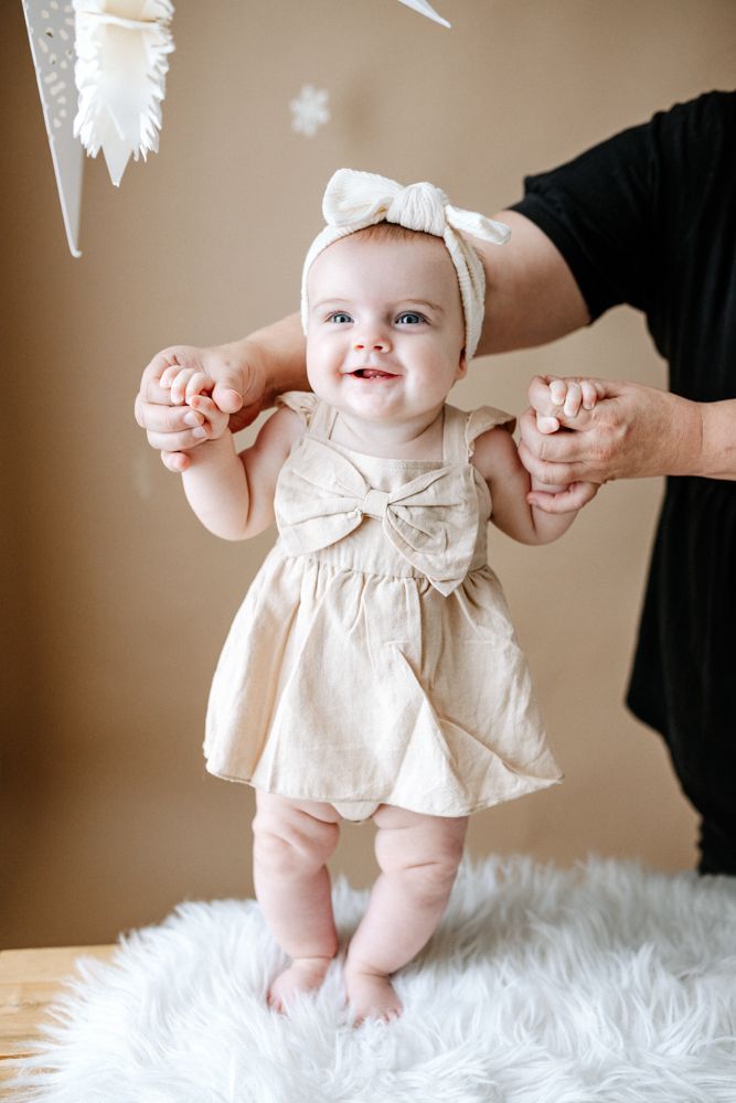 Baby wearing white bow and dress being supported while standing on fluffy white fur surface.