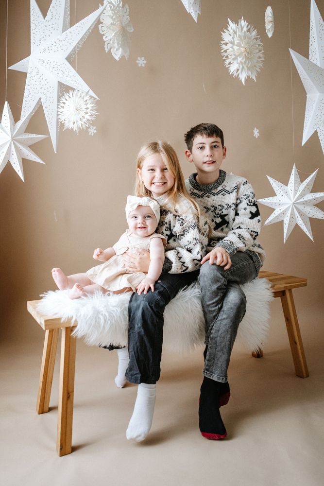 Children sitting on a wooden bench with decorative white paper stars hanging from above in a winter holiday setting.