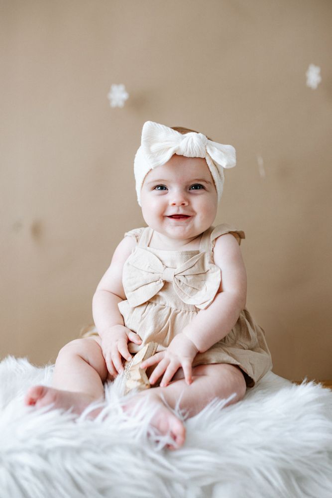 A baby in a white bow headband and beige outfit smiles while lying on soft white fur.